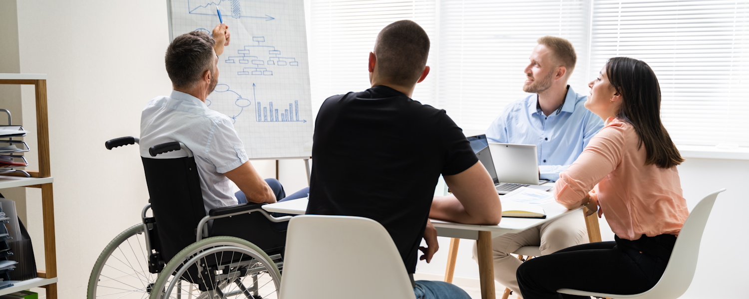 Man in roelstoel legt aan collega's op een whiteboard uit wat een grafiek is.