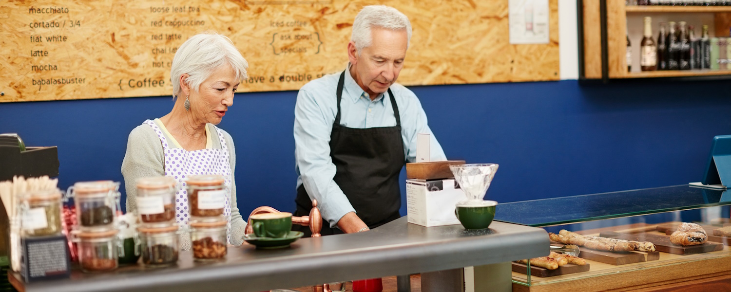 Twee oudere mensen die nog lijken te werken in een cafetaria en staan achter een balie. Beiden willen echter graag pensioen lijkt het