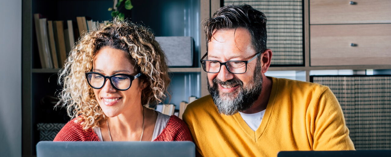 Twee mensen met bril die glimlachend naar hun laptopschermen kijken in woonkamer