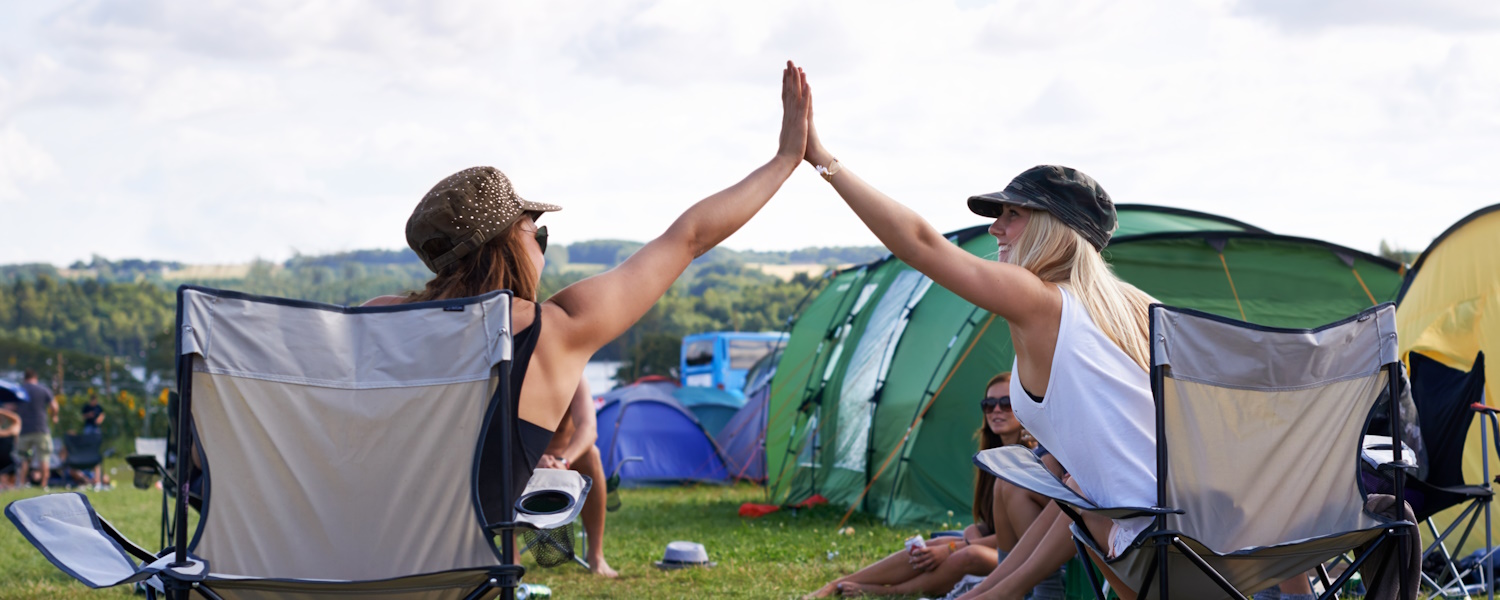 Twee vrouwen met hoofddeksel, zittend in campingstoelen geven elkaar een highfive op de camping