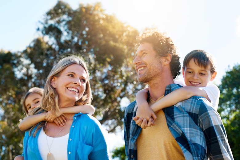 Man en vrouw lopen met kinderen op hun rug, lachend, onbezorgd, in de zon.